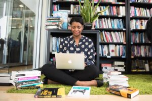 Foto einer farbigen Autorin, die gerade am Laptop etwas schreibt. Sie sitzt auf dem Boden in mitten einer Menge verschiedener IT-Fachbücher. Hinter ihr Bücherregale voller Bücher