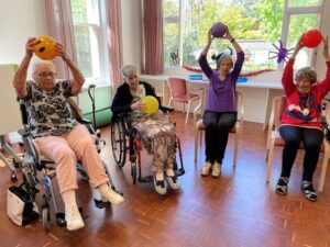 Foto von 4 alten Frauen im Altenheim, die im Rollstuhl oder auf einem Stuhl sitzen und Ballsport machen
